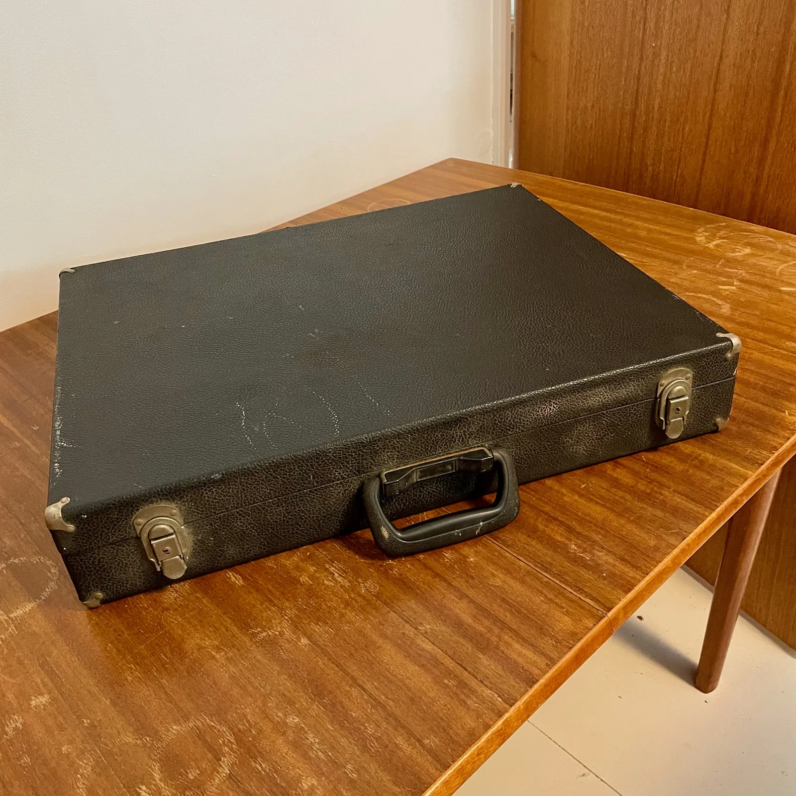 A battered black portfolio case with a plastic handle and metal catches, sitting on a wooden table