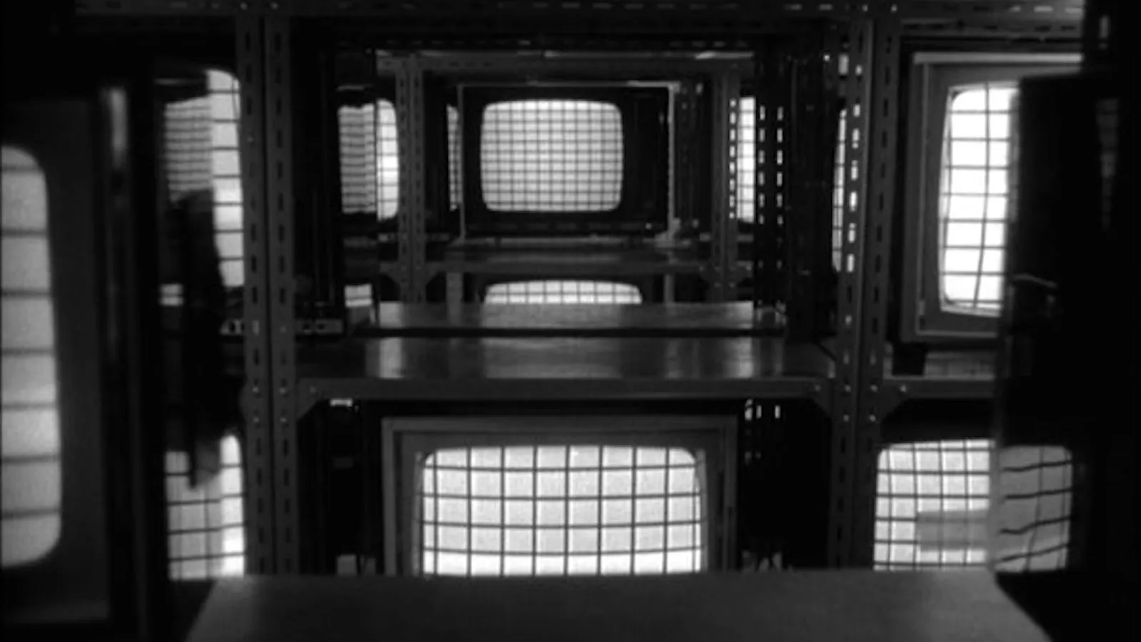 Black and white film still of old television screens stacked on metal shelving, each glowing with a grid pattern